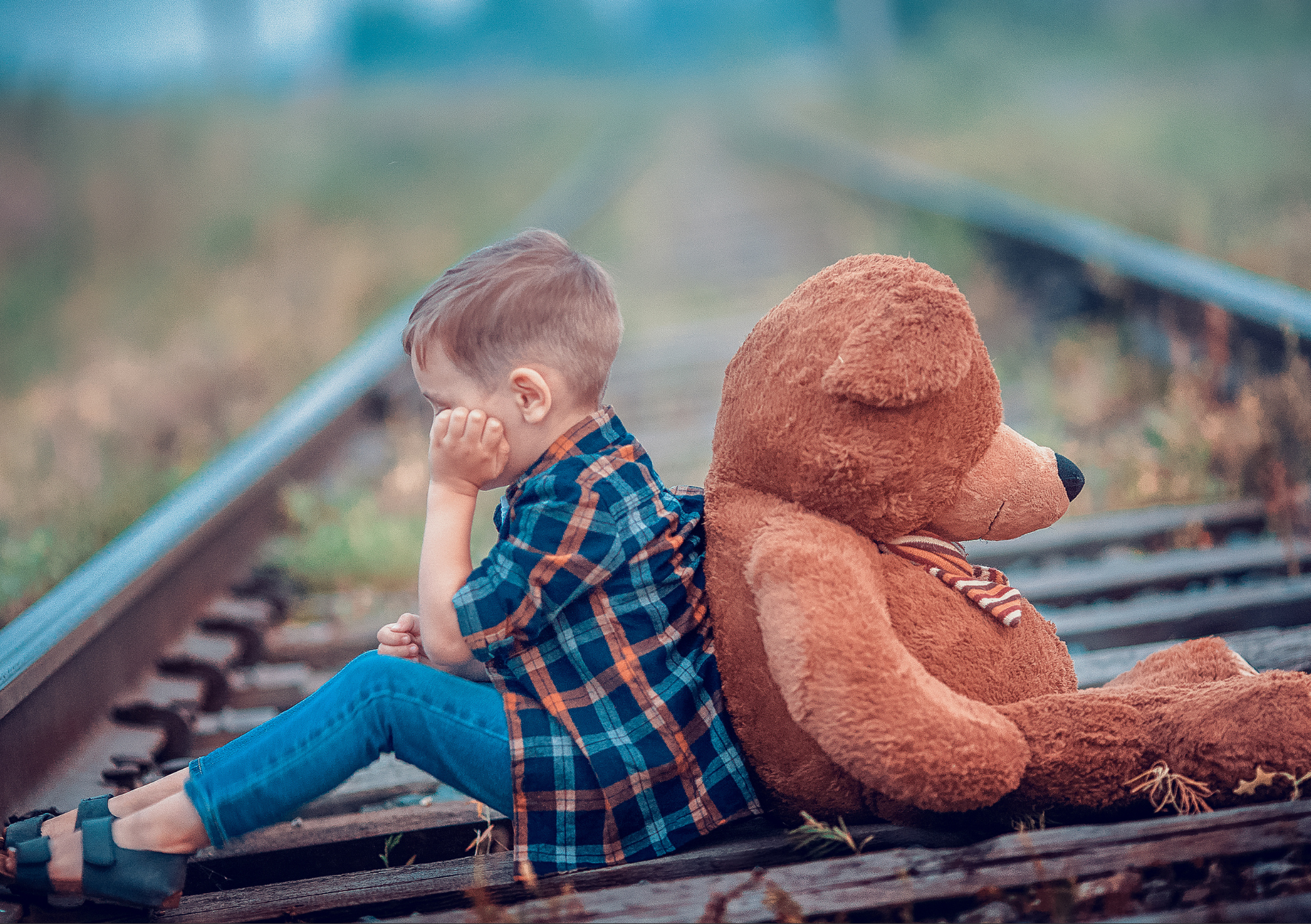 Menino sentado nos trilhos de trem, encostado em um ursinho de pelúcia, com expressão triste ao ar livre.